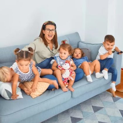 A mother and five young kids laugh and smile while seated together on a lounge room sofa