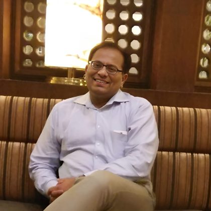 Dr Abubakkar Siddique sits crossed legged on a couch wearing a shirt smiling at the camera